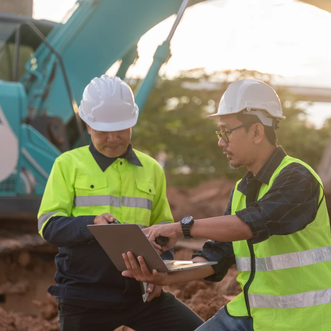 Two engineers in hard hats and high-vis vests reviewing data on a laptop in front of an excavator.
